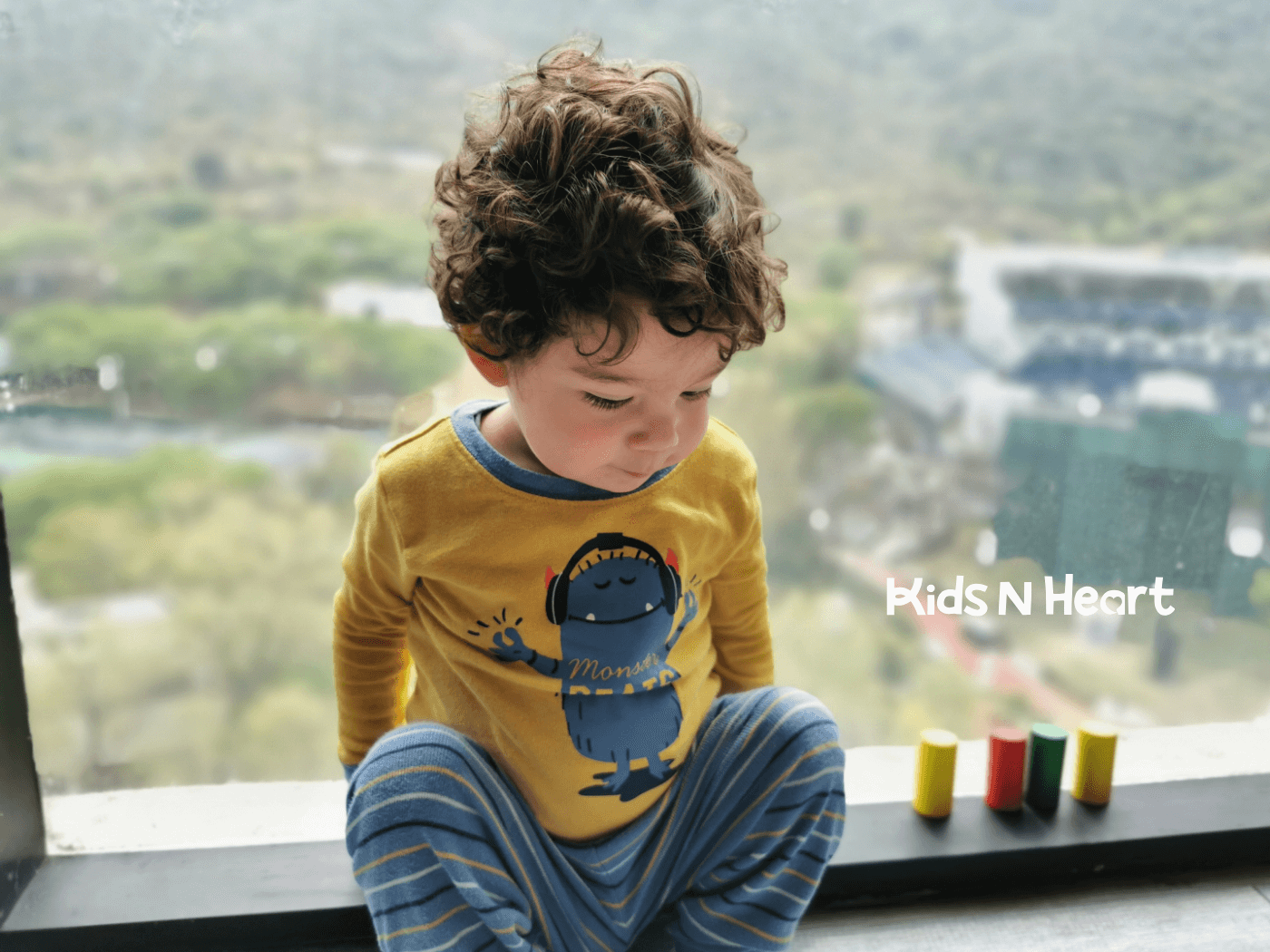 Young child with curly hair sits by a window, calmly focused, with colorful wooden blocks lined up beside them.