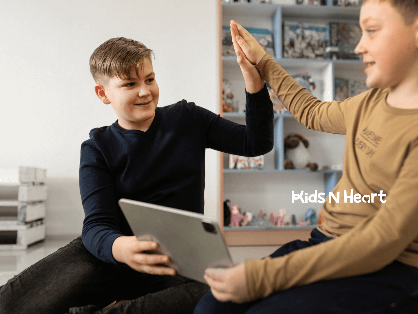Two autistic boys high-five during a learning activity, smiling and sharing a tablet in a supportive, playful indoor setting.