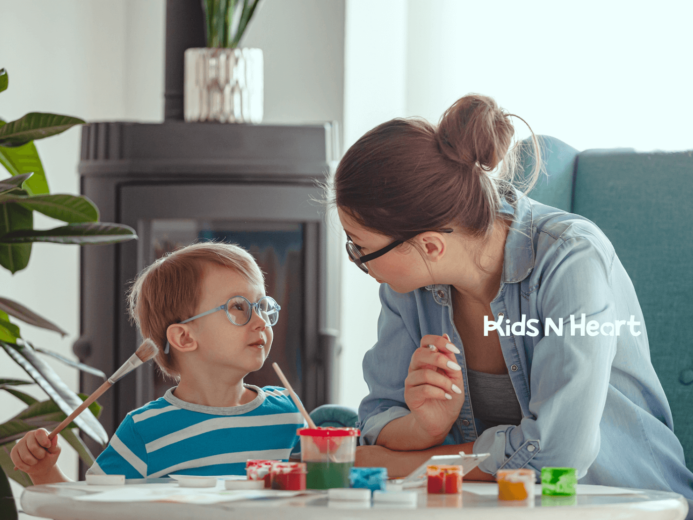 Caregiver and young child with glasses share a warm moment while painting together at a table in a bright, cozy home.