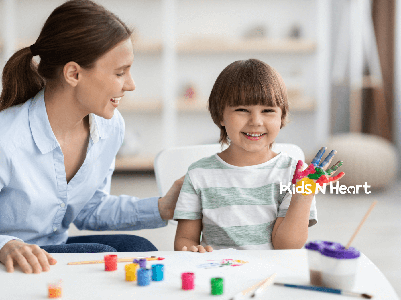 Caregiver smiles beside a young child proudly showing a colorful painted hand during a fun, creative art activity at home.