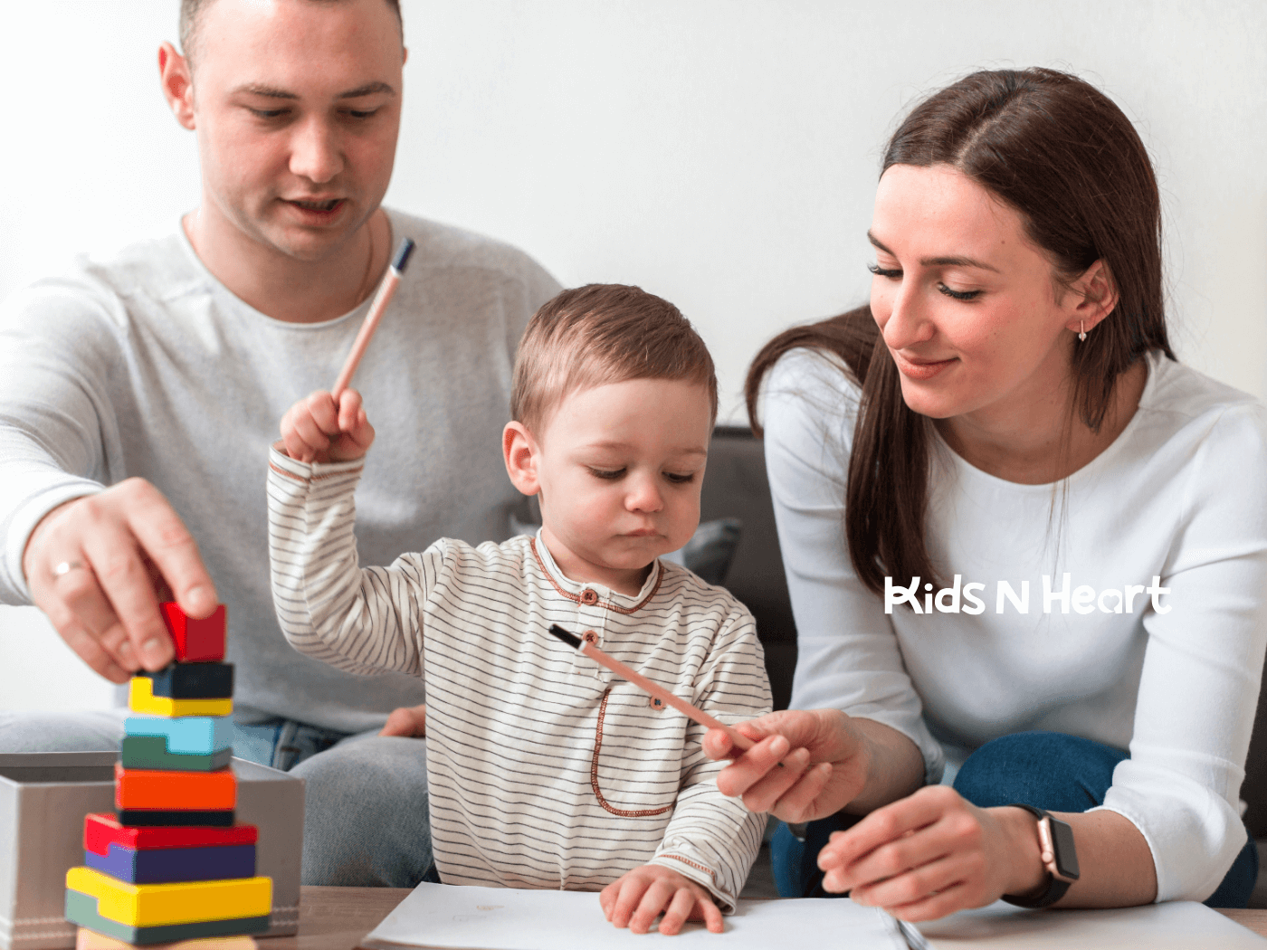 Parents support a toddler stacking colorful blocks and drawing together at home, encouraging learning, focus, and play.