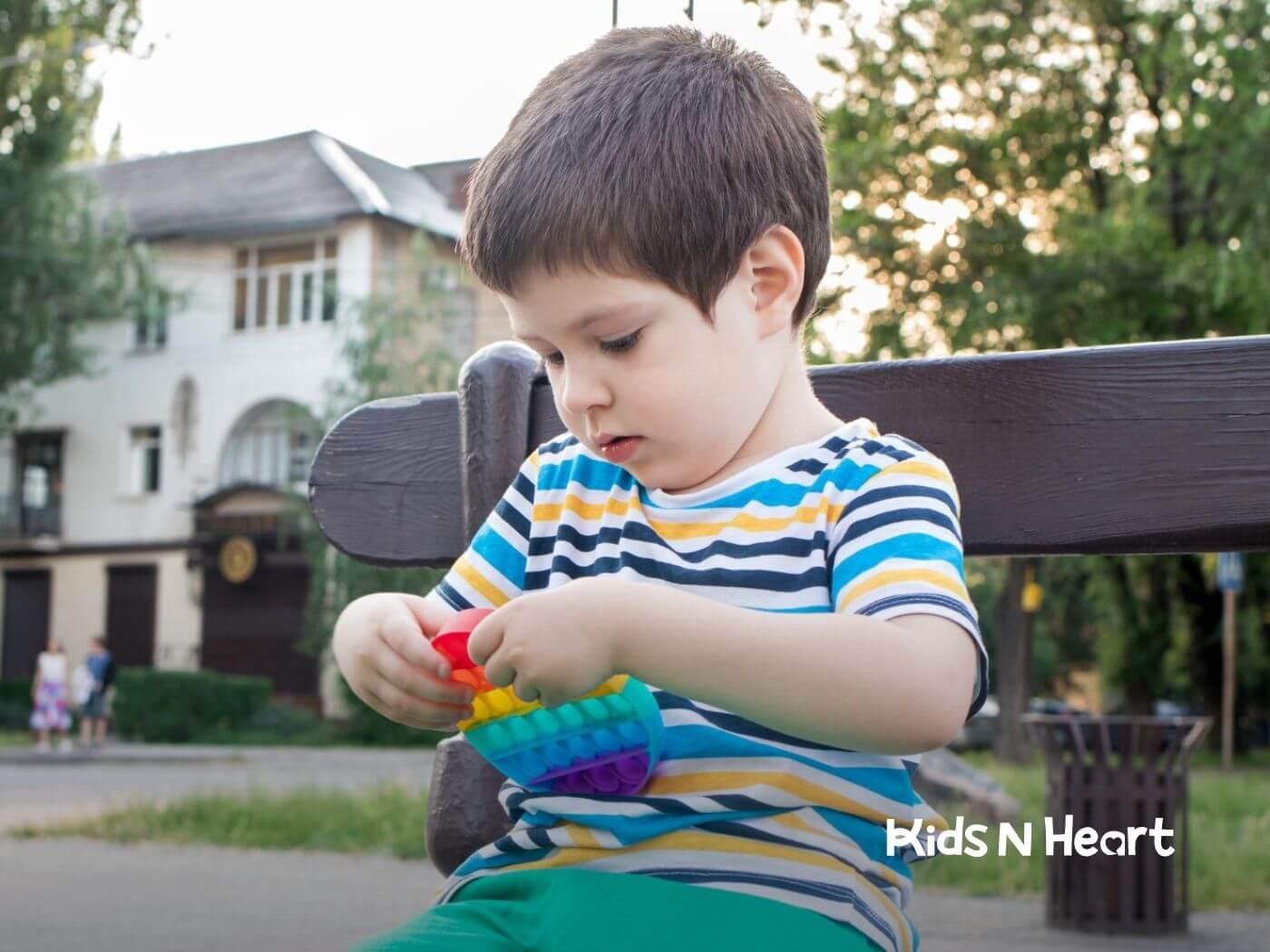 Young boy with autism sitting on a bench playing with a rainbow pop-it toy.