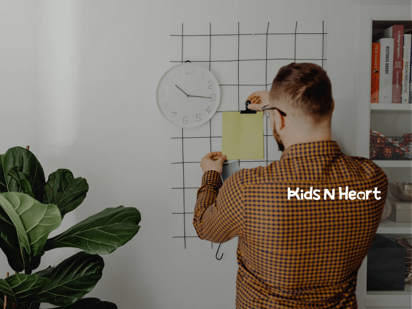 A BCBA in a checkered shirt pinning a note to a grid wall with a clock above it for home-based ABA therapy scheduling.