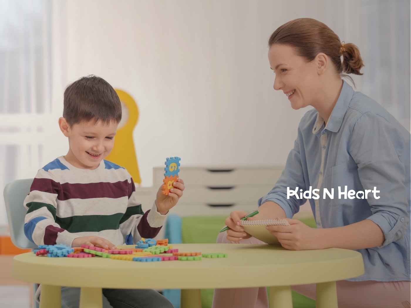 A child and adult happily play with colorful building blocks at a table. The adult takes notes, creating an educational and joyful environment.