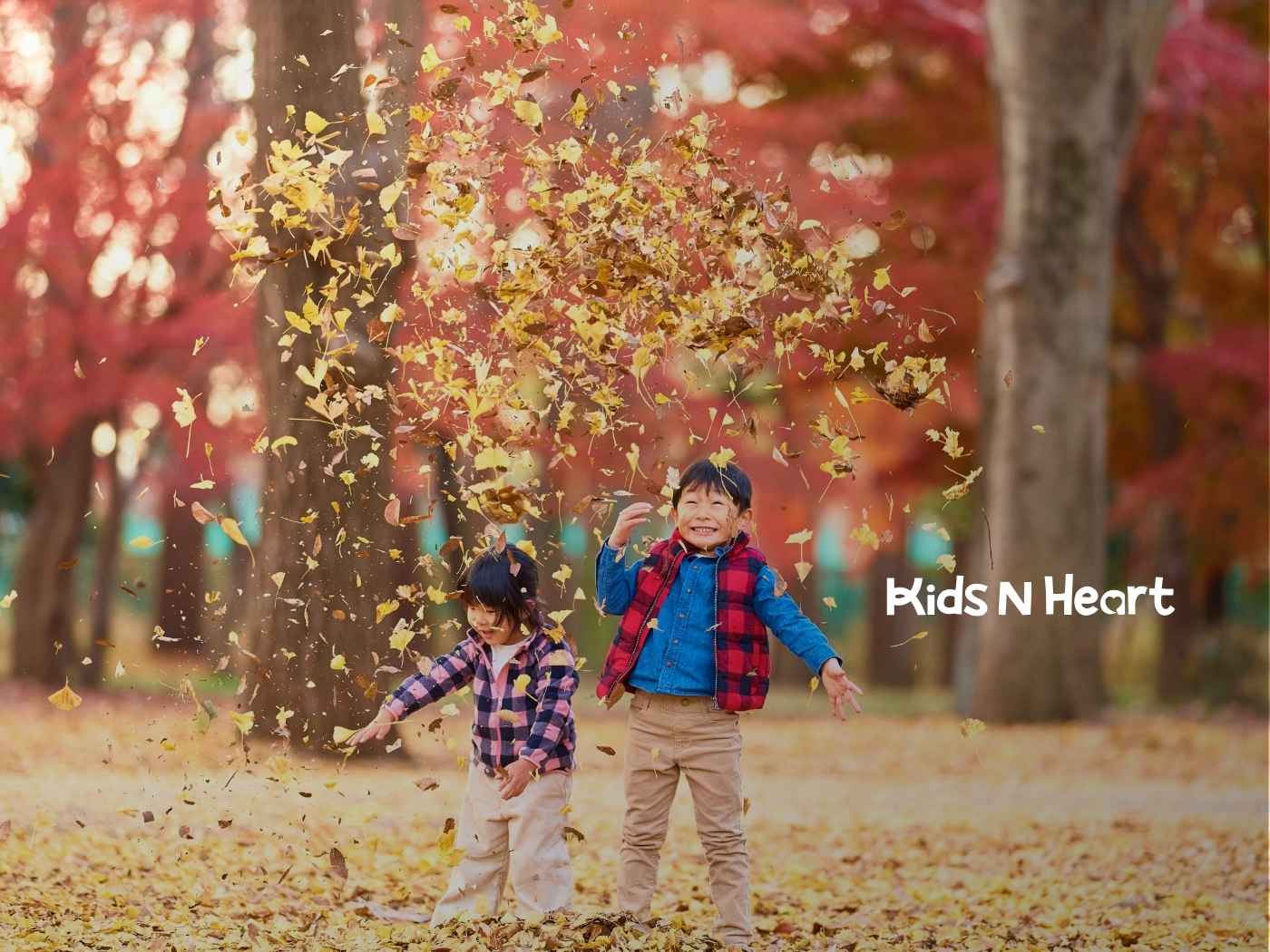 Two children joyfully play with autumn leaves in a park. The background is filled with vibrant red and orange trees, creating a warm, cheerful atmosphere.