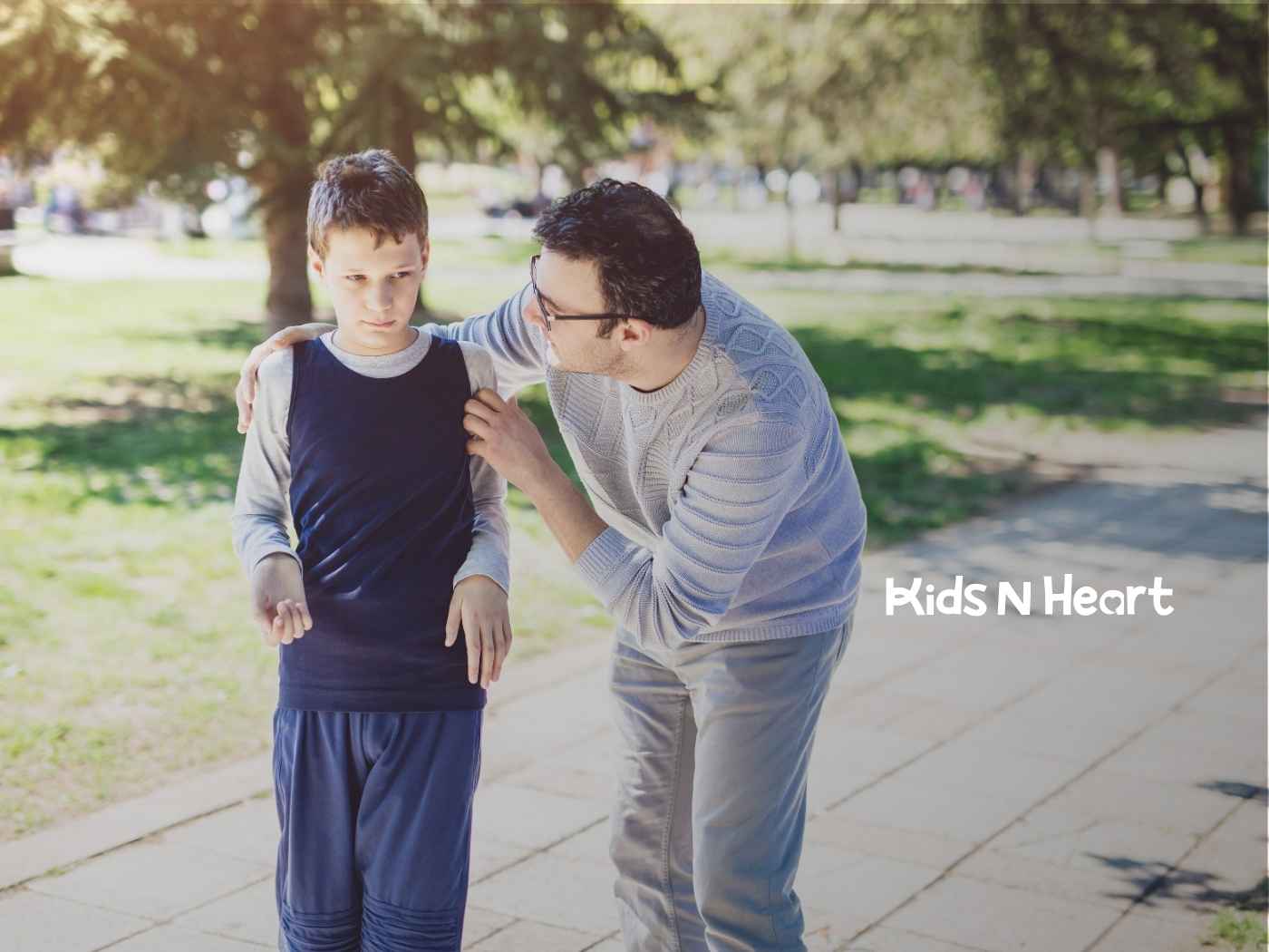 A man gently supports a concerned-looking boy in a park, conveying care. They are surrounded by sunlight, trees, and a paved walkway.
