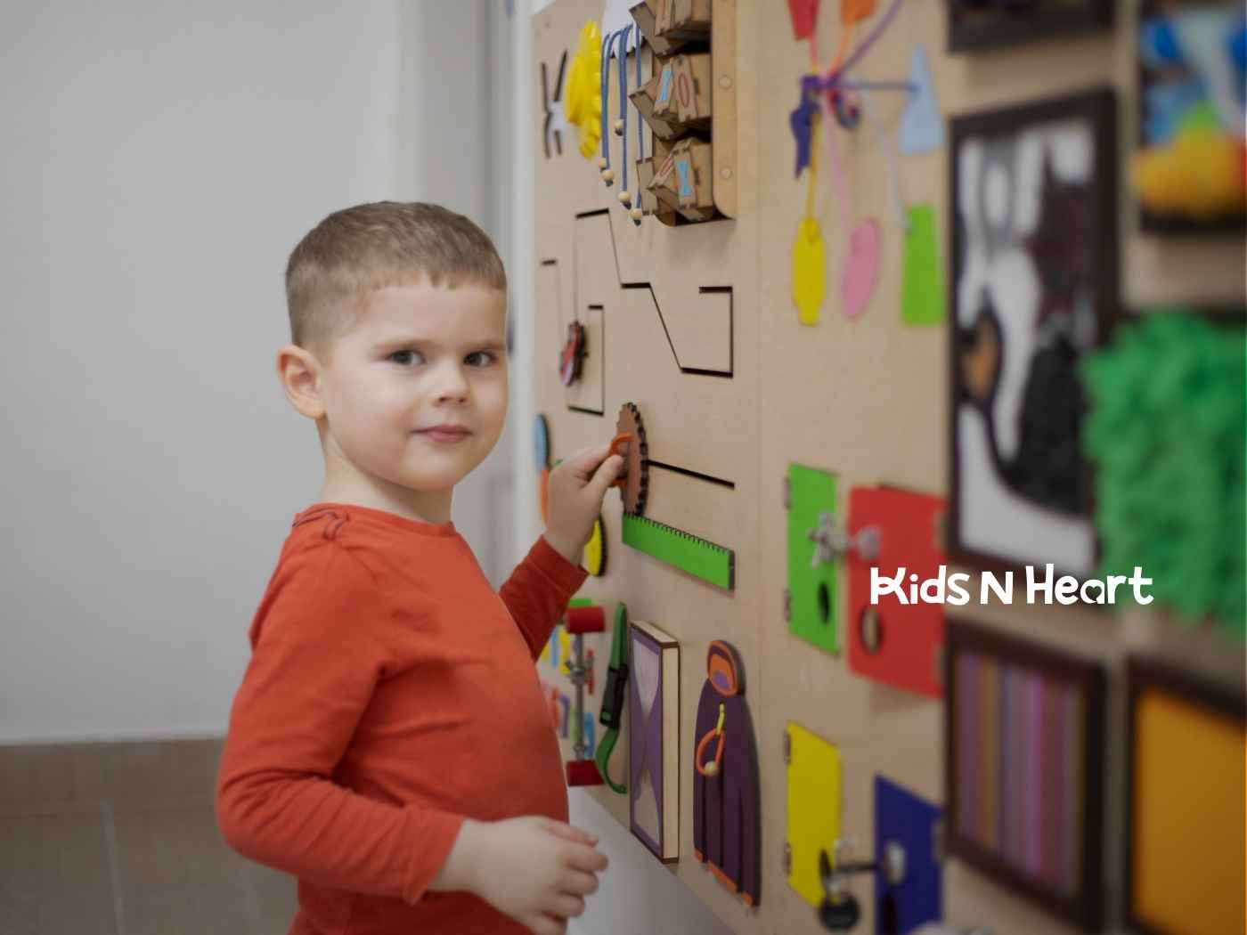 A young child in an orange shirt interacts with a colorful sensory wall full of gadgets and activities, displaying curiosity and focus.