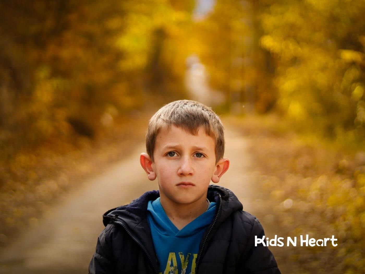 Serious autistic young boy standing on a path surrounded by golden autumn leaves after ABA therapy.