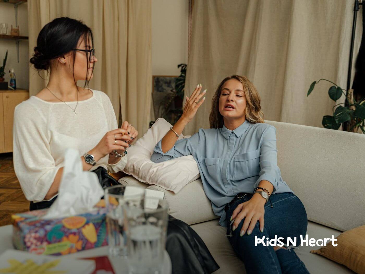 Two autistic women, one is sitting on a couch and the other kneeling on the floor while having an emotional conversation.