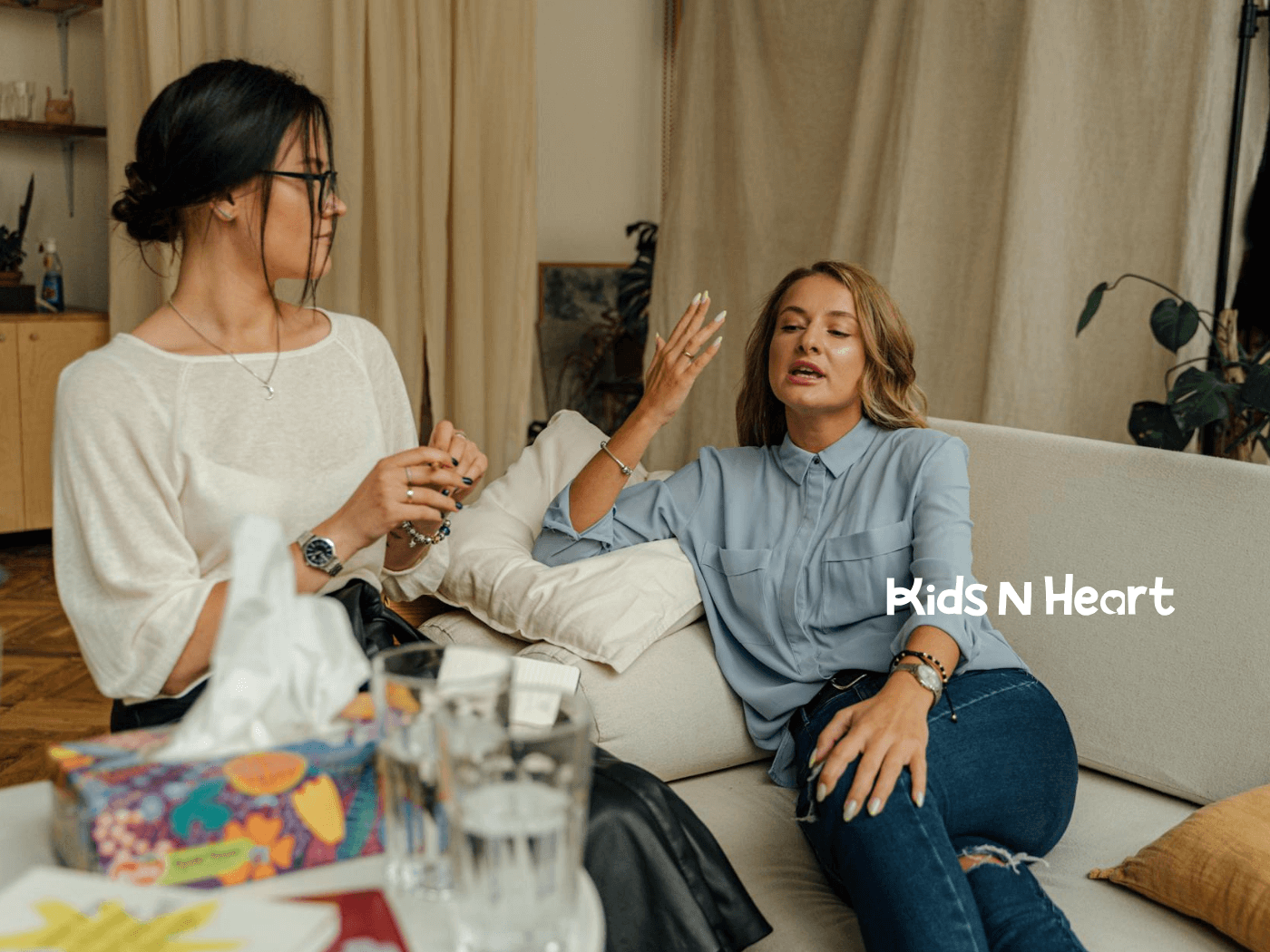 Two autistic women, one is sitting on a couch and the other kneeling on the floor while having an emotional conversation.