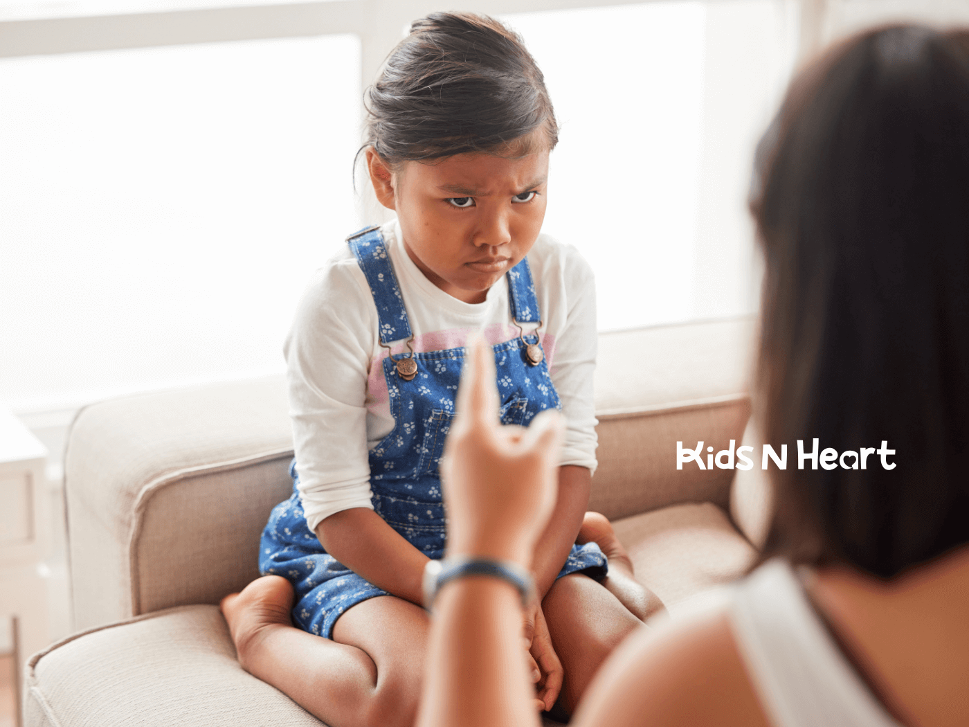 A frustrated autistic young girl sitting on a couch while her mother speaks to her, signaling disapproval.