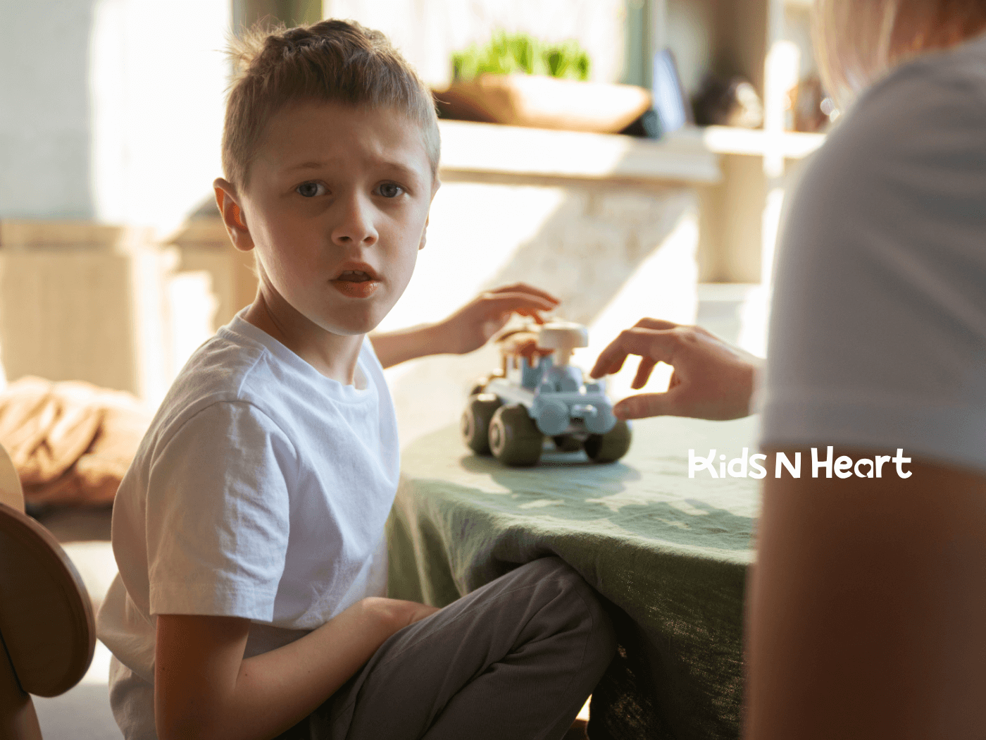 A child sits at a table with a puzzled expression, appearing confused as someone hands them a small toy vehicle.