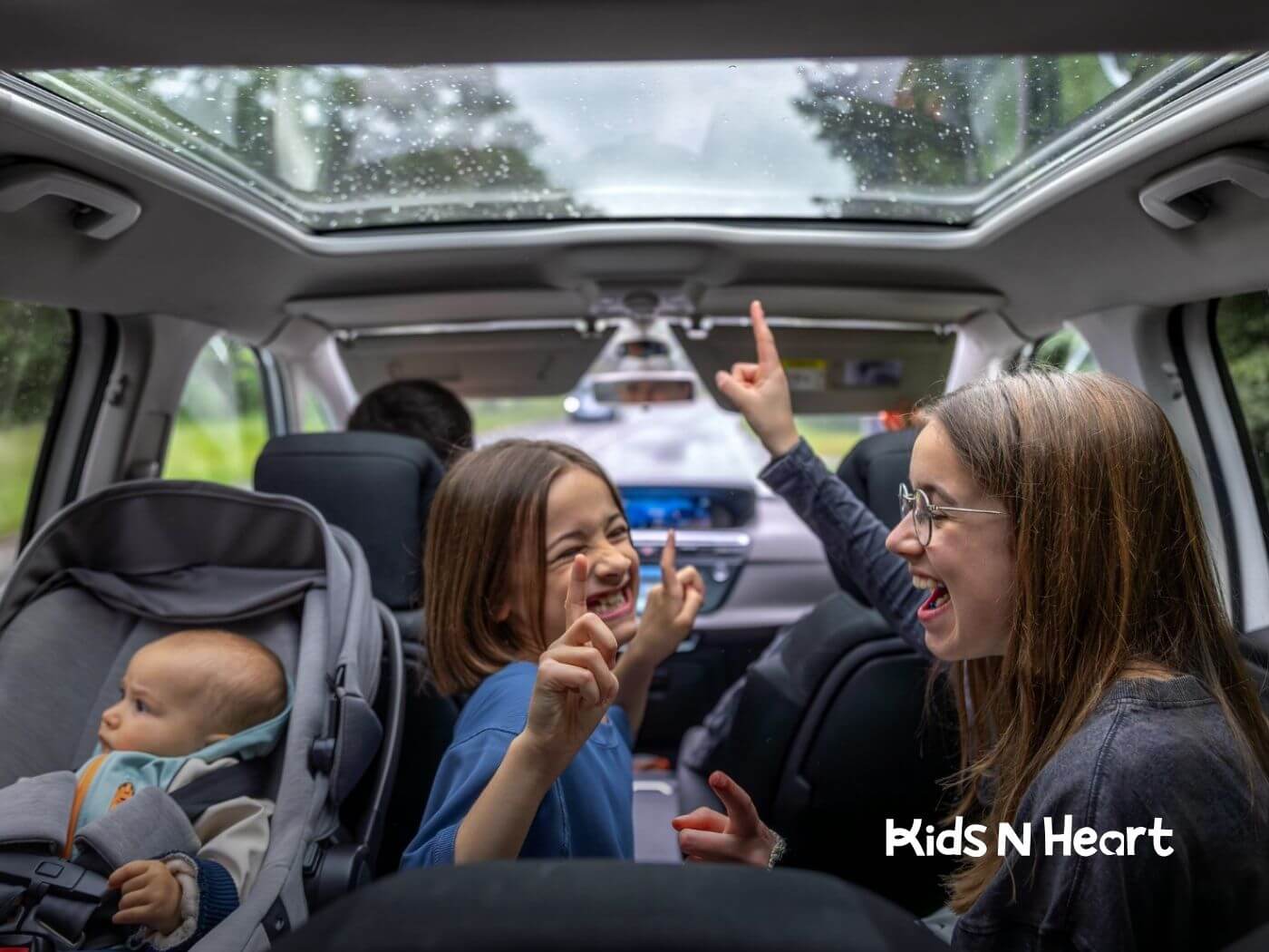 Three children in the back seat of a car, smiling and pointing upward, with a baby in a car seat.