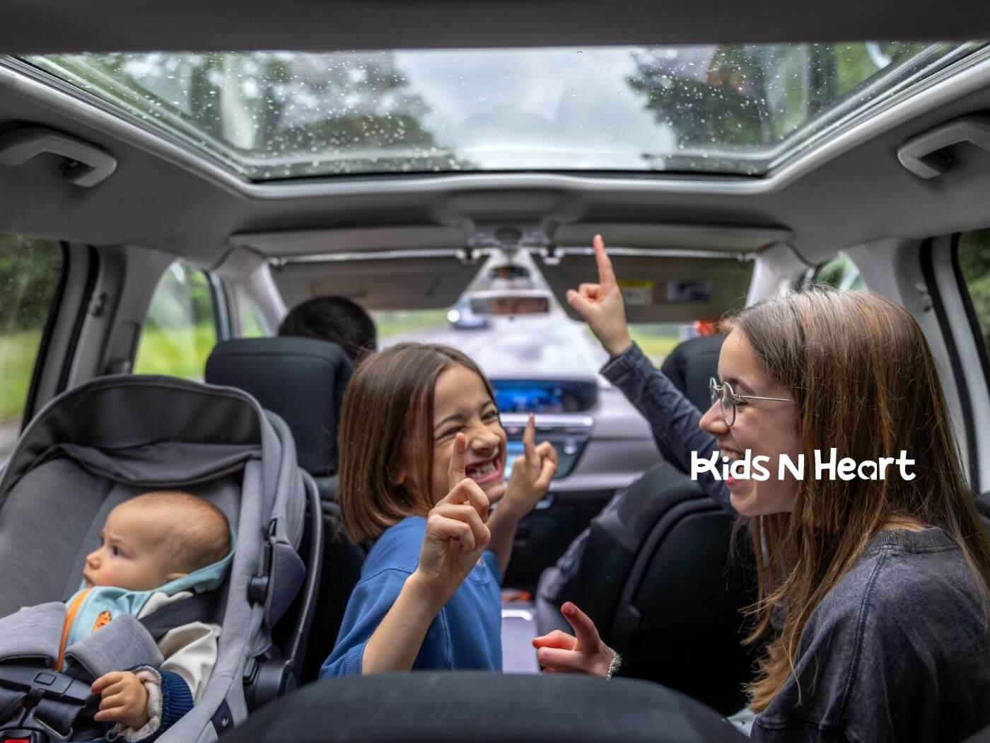 Three children in the back seat of a car, smiling and pointing upward, with a baby in a car seat.